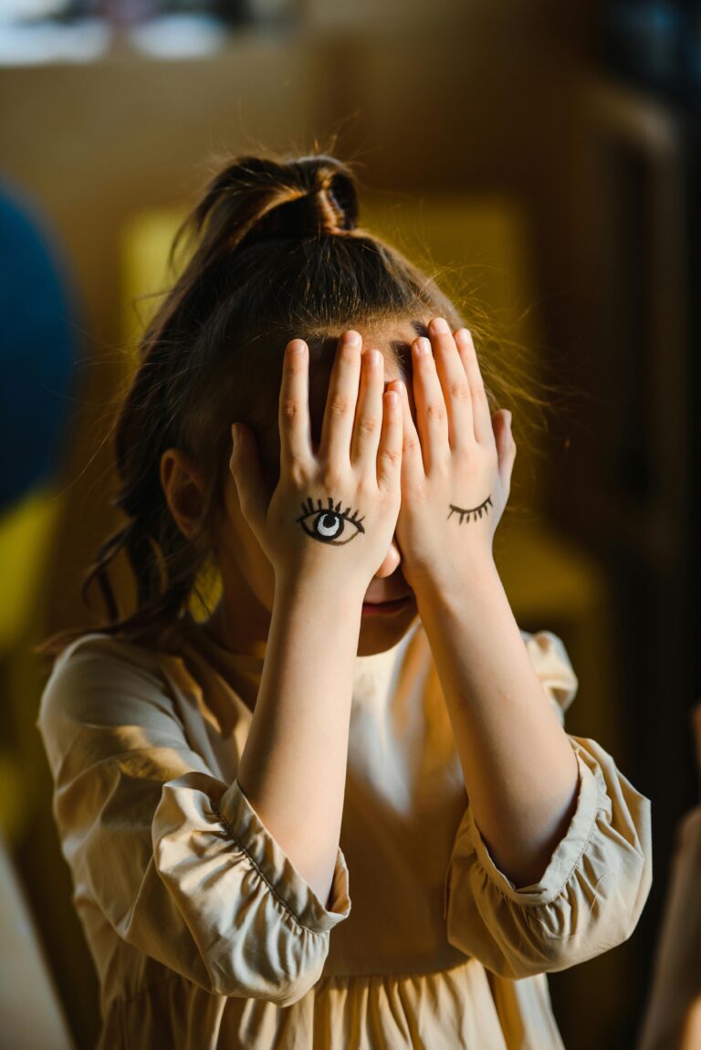 A child with hands covering face, eyes drawn on palms, creating a whimsical image indoors.